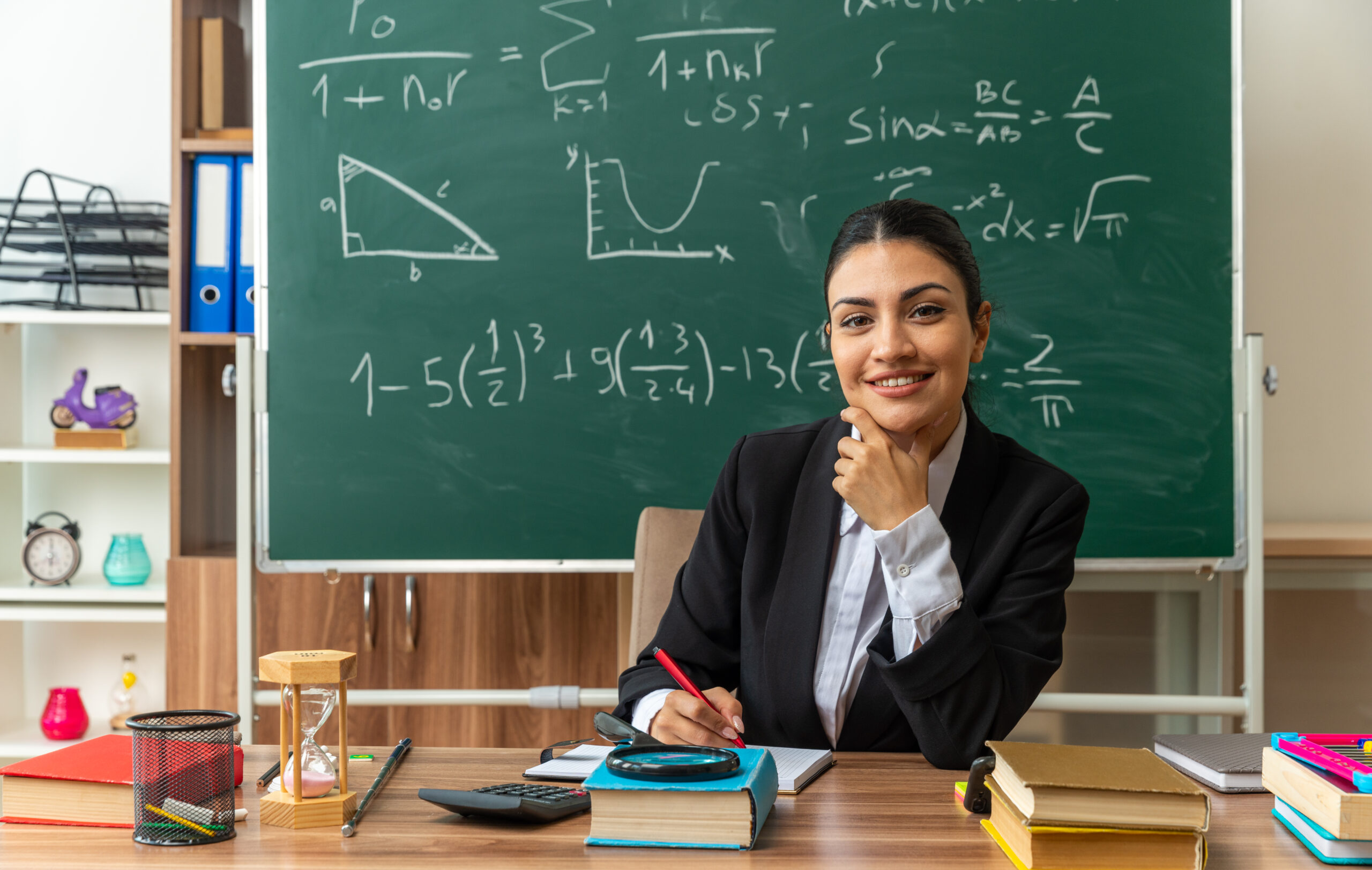 smiling young female teacher sits at table with school tools writing something grabbed chin in classroom